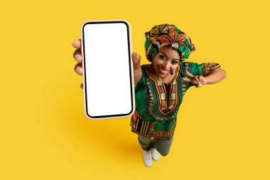 Beyaz beyaz ekranlı, güzel siyah kadın eli ile akıllı telefon, ulusal Afrika kostümü giyen mutlu genç bayan harika bir online anlaşma ve barış jesti gösteriyor, sarı stüdyo arka planı, model, üst görünüm