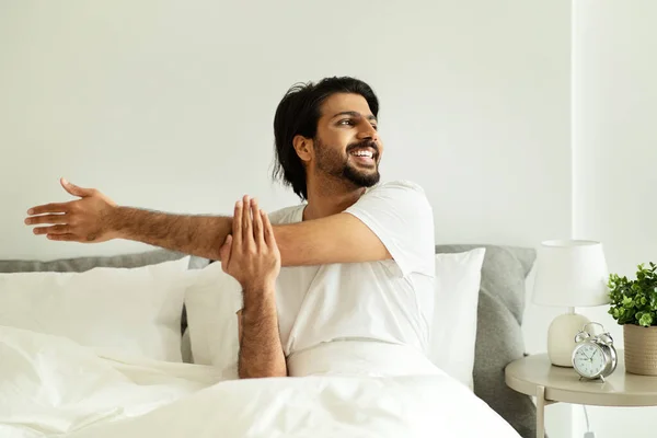 快乐的中东千禧年男人 坐在床上 为双手做伸展运动 为双手伸懒腰 在卧室里享受早晨的愉快 很好的心情 广告和提供 — 图库照片