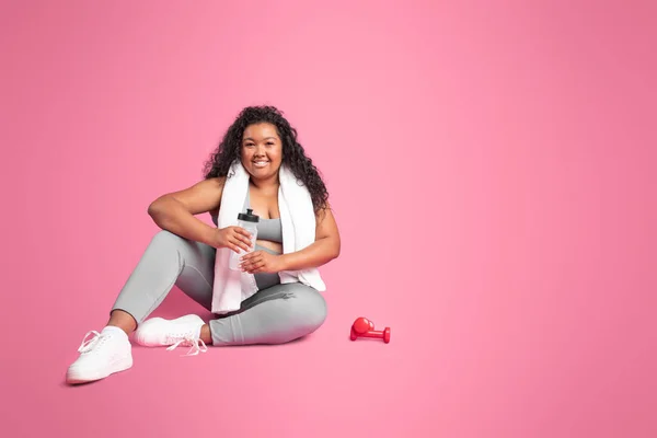 ピンクの背景に休息水のダンベルとボトルとスポーツウェアで幸せなアフリカ系アメリカ人の体の正の女性は コピースペース スポーツ用品 広告やオファー ピラティス フィット感とボディケア — ストック写真
