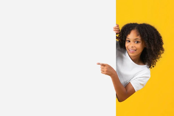 空のスペースで大きなバナーに白いTシャツポイント指で陽気な思春期の黒の巻き少女は 広告をお勧めし 提供し 孤立した黄色のスタジオの背景 生徒の感情 — ストック写真
