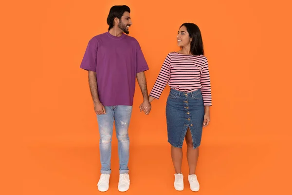 快乐而开朗的笑着的年轻夫妇 他们穿着时髦的休闲装 手牵着手 面面相觑 在橙色的工作室背景下合影 — 图库照片