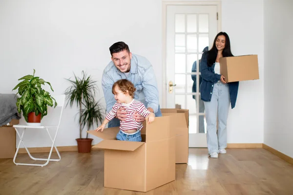 Happy Family Riding Their Son In Cardboard Box While On Moving Day, Young Parents And Cute Toddler Child Relocating To Their New Home, Having Fun Together While Unpacking Things, Free Space