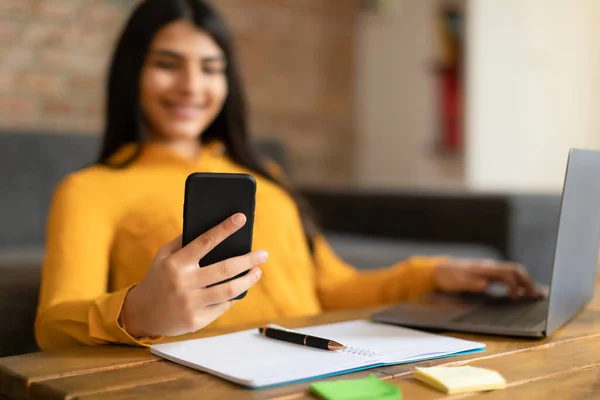 年轻的西班牙女士与笔记本电脑坐在桌旁 一边用手机 一边微笑着在网上购物 一边浏览现代网上银行应用软件 一边精挑细选地进行特写 — 图库照片
