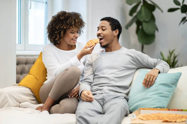 快乐美丽的千年黑人情人坐在沙发上 吃披萨 妻子喂丈夫 爱吃非洲裔美国男人和女人在家里共度周末 复制空间 — 图库照片