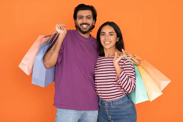 黑色星期五 季节性销售 快乐的东方年轻夫妇身着休闲装拥抱在一起 摆出一副五颜六色的购物袋 与橙色工作室隔离在一起 — 图库照片