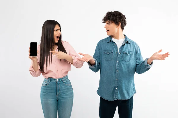 Digital Problem. Angry Girlfriend Showing Phone With Blank Screen To Confused Boyfriend, Discontented With Bad Mobile Offer Posing On White Background. Conflicts And Internet Haters In Social Media