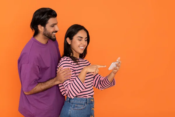 オレンジ色のスタジオの背景に一緒にスマートフォンを使用して楽しい魅力的な若いインドの愛好家 幸せな東部の男性と女性でカジュアル素敵なコンテンツを見てオンラインと笑顔 コピースペース — ストック写真