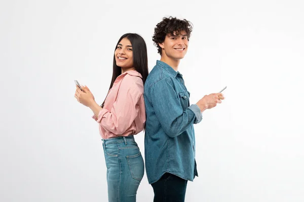 ソーシャルメディアアプリケーション スマートフォンを使用して若いカップルを笑顔立ちバックホワイトスタジオの背景に立ちます 携帯電話の広告のモバイルオファーを介して若者のテキストとサーフィンのウェブ — ストック写真
