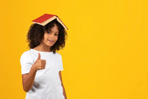 頭の上の本が付いている白いTシャツの中のアフリカ系アメリカ人の少女の笑顔は 黄色のスタジオの背景に隔離された親指を示しています 推奨読書 オファー — ストック写真