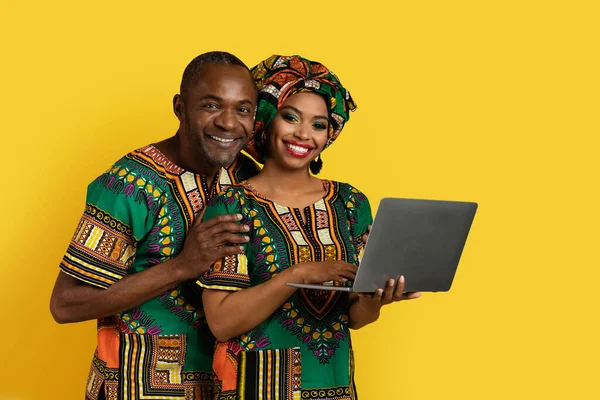 幸せな美しい黒カップル身に着けている国内のアフリカの衣装Websafing上のラップトップPc上で黄色のスタジオの背景 抱擁と笑顔でカメラ 愛の絆 コピースペース — ストック写真