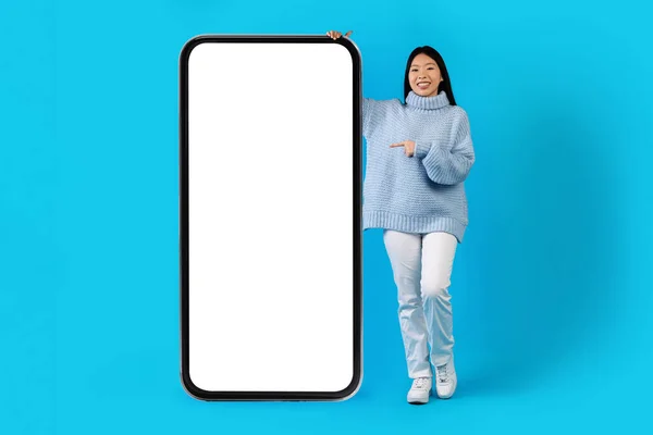 提供移动应用程序 快乐而美丽的年轻中国女士 穿着暖和的毛衣 用白色的空屏幕对着巨大的手机 模仿蓝色的工作室背景 复制空间 — 图库照片