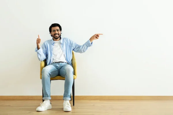 积极的中东千禧年男子展示了大拇指 指指点点空旷的空间 坐在扶手椅上的白墙背景 广告和报盘 — 图库照片