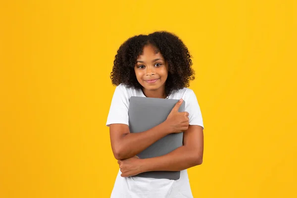 stock image Happy adolescent curly black girl in white t-shirt hugs tablet, isolated on yellow studio background. Device for blog and website for education, study, love to social networks and chat