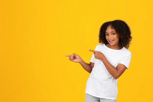 穿着白色T恤的年轻的卷曲非洲裔美国女孩微笑着 手指指向空旷的空间 推荐销售 广告和提供 孤立的黄色工作室背景 学生情绪 — 图库照片