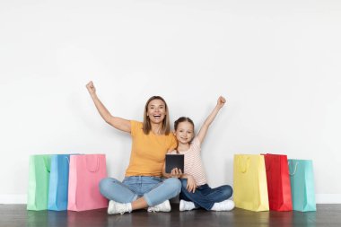 Büyük satışlar. Dijital tabletli mutlu anne ve küçük kızı beyaz duvarın yanında duran alışveriş torbaları, sallanan yumruklar, neşeli anne ve çocuk internetten alışveriş yapıyor.