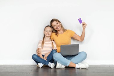 Kolay ödemeler. Tatlı küçük kız ve dizüstü bilgisayarı ve kredi kartıyla beyaz duvarın yanında oturan mutlu annesi, gülümseyen milenyum kadını ve E-ticaretten hoşlanan kadın çocuğu, kopyalama alanı.