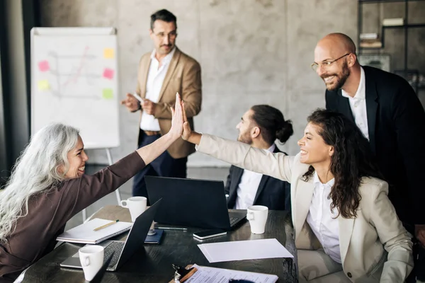 Joy Of Business Success. Joyful Team Of Coworkers Giving High Five During Corporate Meeting, Celebrating Successful Teamwork Sitting At Table In Modern Office Interior. Selective Focus