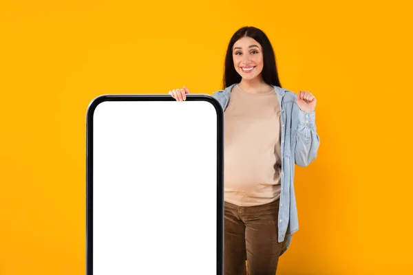 素晴らしい提案だ 白い画面と黄色の背景 モックアップの上にはいジェスチャーで大きなスマートフォンの近くに立って幸せな若い妊婦 妊娠のアプリやウェブサイトをお勧めする期待の女性 — ストック写真
