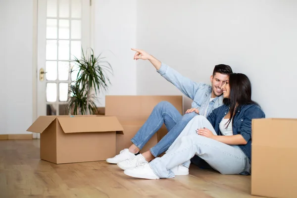 Young Happy Couple Planning Interior Design In Their New Home After Moving, Smiling European Spouses Sitting On Floor Among Cardboard Boxes And Pointing Away, Imagining Decorations And Furniture