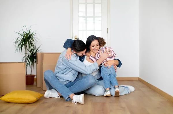 New Beginnings. Happy Family Of Three Embracing At Home On Moving Day, Cheerful Young Parents And Child Hugging While Sitting On Floor Among Cardboard Boxes, Celebrting Relocation To New Flat