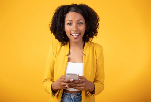 携帯電話を使用し カメラで笑みを浮かべて忙しい髪を持つ幸せな黒の女性の肖像画は 黄色のスタジオの背景に孤立立っている アフリカ系アメリカ人女性がオンラインでチャットしたり ソーシャルメディアを閲覧したり — ストック写真