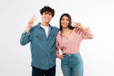 Sevgi ve Barış. Beyaz Stüdyo Arkaplanının Üzerinde Duran Kameraya Doğru Hareketli Zafer İşareti Veren Mutlu Genç Çift. Neşeli arkadaşlar kucaklaşıp V-İşareti Sembolü gösteriyorlar
