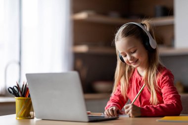 Uzaktan eğitim. Kulaklıkla gülümseyen, laptopla çalışan ve notlar alan, evde masa başında oturan, internetten ders alan, not alan, boşluğu kopyalayan küçük bir kız çocuğu.