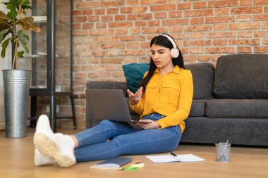 Kablosuz kulaklıklı neşeli İspanyol kadın dizüstü bilgisayarda video görüşmesi yapıyor, evden uzaktan ders alıyor, web kamerasına konuşuyor ve el kol hareketi yapıyor.