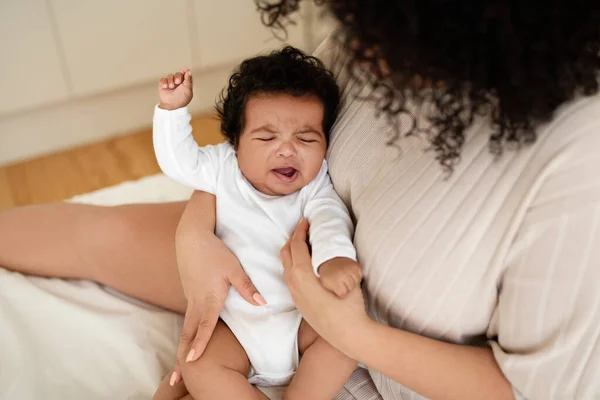 Black millennial mom calms crying sad little baby on white bed in bedroom. Disease treatment, teeth growing, child care, parenthood and family at home, health care and love