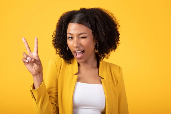 快乐的黑卷曲女人眨了眨眼 手拿着和平标志 在黄色的背景下摆出孤立的姿势 拍了下来 广告和服务 时尚博客 美容护理 积极的生活方式 — 图库照片