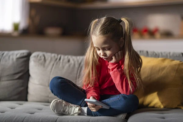 stock image Upset Little Girl Using Mobile Phone While Relaxing On Couch At Home, Bored Preteen Female Child Playing Games Or Browsing Internet On Smartphone, Sad Kid Checking New App, Copy Space