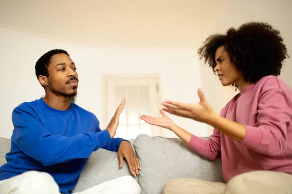 Relationship And Conflicts. African American Spouses Quarreling Sitting On Sofa, Husband Gesturing Stop To Calm Down Emotional Wife At Home. Couple Arguing Suffering From Marital Crisis