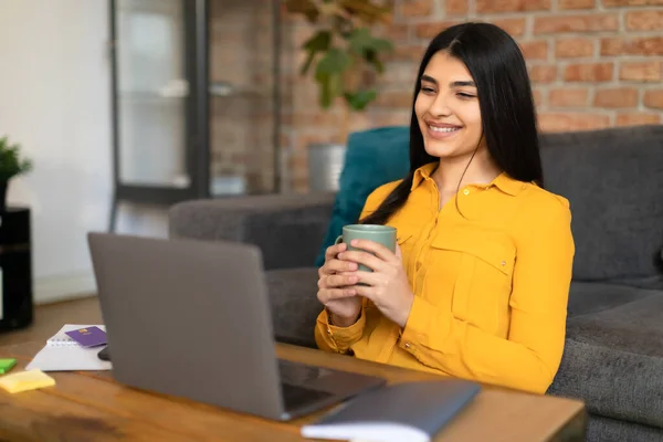 ラップトップでビデオを見ている陽気なスペイン人女性と自宅でコーヒーを飲んで 幸せな女性はコンピュータ画面を見て オンラインクラスの後に休んで スペースをコピー — ストック写真