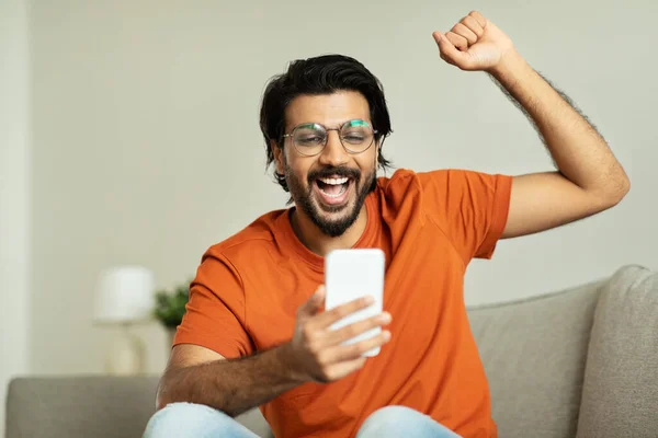 Glad excited millennial middle eastern man in glasses rejoices to victory, celebrating success, raises hand up, looks at smartphone, sits on sofa in living room interior. Great news, ad and offer