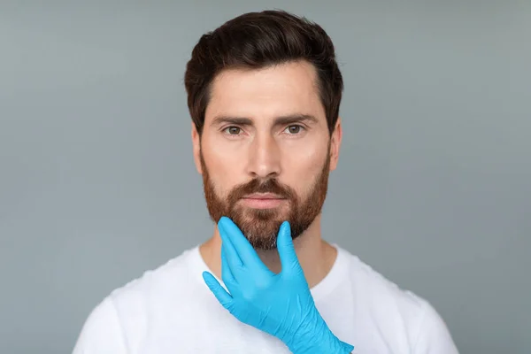 専門家の話だ 灰色のスタジオの背景に中年男性の顔に触れる保護医療用手袋で美容師の手 整形手術と審美的な美容コンセプト — ストック写真