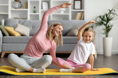 Mutlu bir genç anne ve küçük kızı evde spor yapıyor neşeli bir anne ve tatlı bir kız çocuğu yan eğilme egzersizleri yapıyor oturma odasının iç kısmında spor yapıyor.