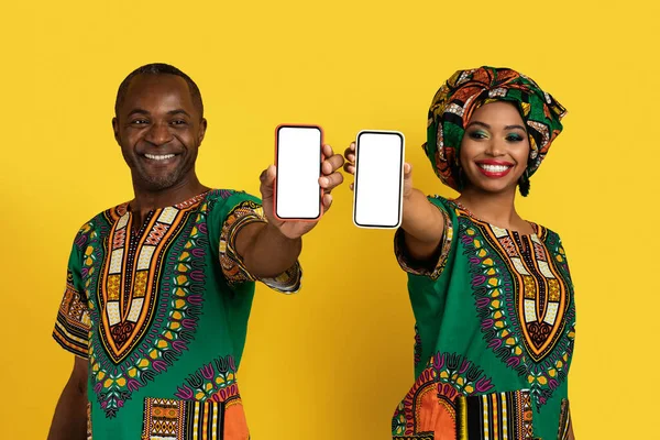 白い空の画面で携帯電話を示すアフリカの衣装で美しい笑顔の黒い恋人の男と女 楽しいモバイルアプリをお勧めします 黄色のスタジオの背景 コピースペース — ストック写真