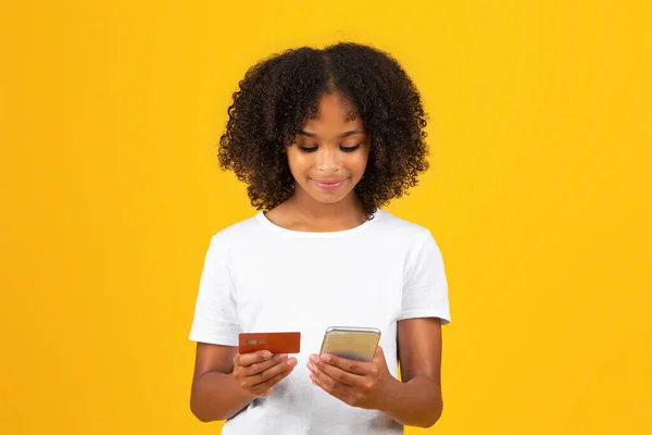 白いTシャツを着た幸せな思春期の巻き黒の女の子は 黄色のスタジオの背景に隔離されたオンラインショッピングのためにクレジットカードとスマートフォンを使用しています 販売のためのデバイス 金融銀行アプリ — ストック写真