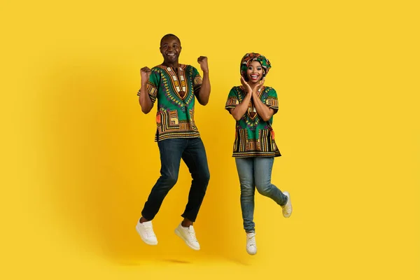 素晴らしいニュース 運の概念 明るい民族衣装を身に着けている幸せなアフリカ系アメリカ人のカップルがジャンプアップし 黄色のスタジオの背景にジェスチャー フル長さ コピースペース — ストック写真