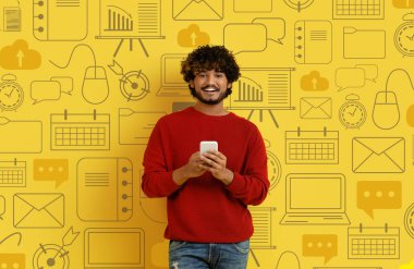 Sarı arka planda çeşitli dijital dünya simgelerinin üzerine telefonla poz veren sıradan, mutlu, yakışıklı Hindu bir adam. Modern teknoloji kavramı, mobil uygulamalar, sohbet gpt kavramı