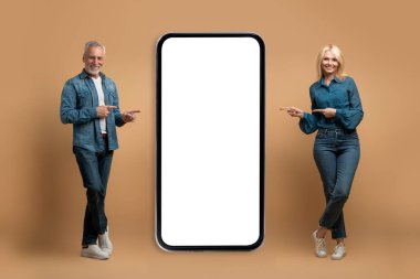 Heyecan verici online teklif, eğlence için güzel mobil uygulama. Neşeli yaşlı bir çift, günlük kot giyip beyaz ekranlı, renkli arka planlı, taklit edilmiş büyük akıllı telefonu işaret ediyorlar.