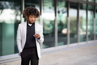 Siyah erkek, orta yaşlı bir çalışan cep telefonu kullanıyor, arkadaşlarıyla sohbet ediyor ya da ara verirken internette yeni iş fırsatları arıyor, ofis binasının yanında yürüyor, boşluk kopyalıyor.