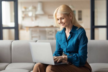 Diz üstü bilgisayar kullanan ve gülümseyen mutlu Avrupalı kadın, evdeki koltukta oturuyor, internette çalışıyor ya da internet hayatına takılıyor, boş yer arıyor. Gadgets teknolojisi