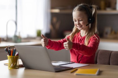 Çevrimiçi Ders. Ev içinde masa başında otururken dizüstü bilgisayar kullanan ve başparmaklarını Webcamera 'da gösteren tatlı liseli kız, uzaktan öğrenmenin, kopyalamanın, boşluğun tadını çıkaran mutlu çocuk.