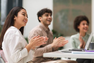 İş Başarı Kutlaması. Mutlu çok kültürlü meslektaşlar el çırpıyor, başarıyı kutluyor, masada oturuyor ve ofise yan bakıyor. Asyalı İş Kadınına Seçici Odaklanma