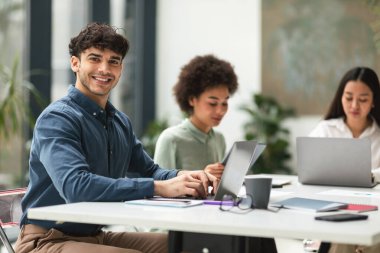 Şirket Başarı. Mutlu Arap iş adamı, iş yerinde farklı iş arkadaşlarının yanında dizüstü bilgisayar kullanıyor, modern ofis içi toplantılarında güvenle gülümsüyor.
