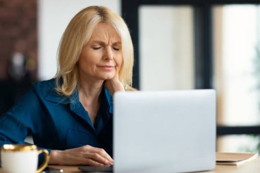 Evde dizüstü bilgisayarla çalışırken boyun spazmından muzdarip olgun bir kadın masasında oturup kaslarını ovuyor. İş kadınının boynuna rahatsız edici bir durum yüzünden kramp giriyor.