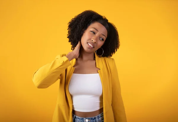 Despaired sad black curly woman suffering from neck pain, making massage, standing on yellow background. Health problems, muscle pain after training, overwork and arthritis, injury