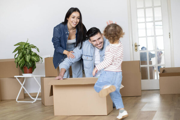 Cheerful Family Of Three Having Fun While Relocating Home, Happy Young Parents And Their Toddler Son Fooling Together While Unpacking Things On Moving Day, Playing With Cardboard Boxes In Living Room