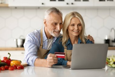 Mutfakta dizüstü bilgisayar ve kredi kartı kullanan, gülümseyen olgun eşler yiyecek siparişi veren, internetten alışveriş yapan, evdeki bilgisayardan alışveriş yapan,